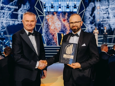 Vidutinio plano kadras, kuriame matyti du vyrai, apsirengę oficialiais vakariniais kostiumais, spaudžiantys vienas kitam rankas. Dešinėje esantis vyras, Maciej Góralski, laiko įrėmintą apdovanojimą su auksine emblema, iš kurios matyti, kad tai „Entrepreneur of the Year 2022“ apdovanojimas. Neryškiame fone matoma scena su abstrakčiomis geometrinėmis struktūromis ir dinamiškais mėlynos bei raudonai oranžinės spalvos grafiniais raštais. Prislopinto apšvietimo renginių salėje prie stalų sėdi svečiai.