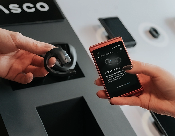Vasco Translator E1 ausinės ir Vasco Translator Q1 rodomos kartu; Q1 ekrane rodomas ausinių poravimo sąsaja, o fone matomas Vasco logotipas.