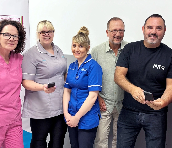 Penki besišypsantys žmonės stovi kartu, apsirengę sveikatos priežiūros drabužiais, du iš jų laiko vertimo įrenginį.