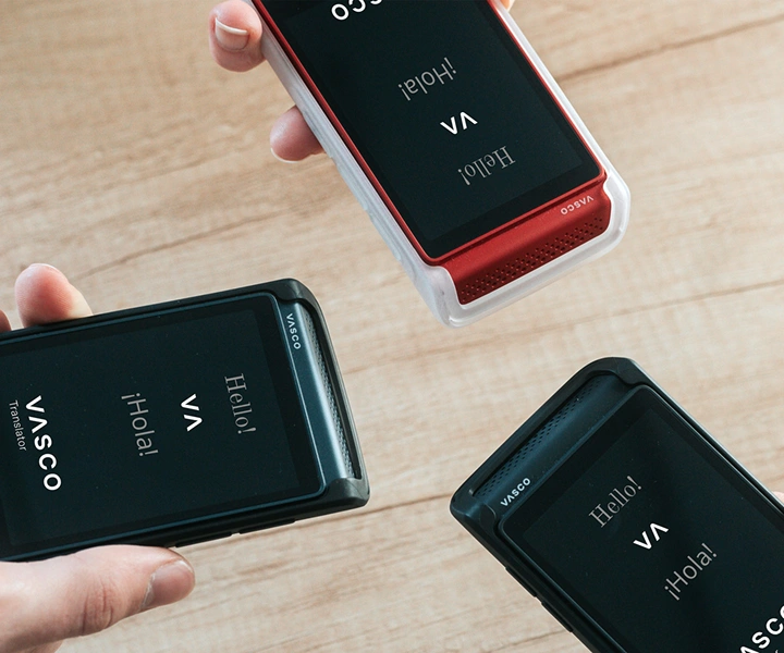 Trys žmonės, laikantys Vasco Translator Q1 įrenginius, sujungia juos; ekranuose matomas pradinis ekranas su Vasco logotipu ir užrašais „Hello!“ bei „¡Hola!“. Įrenginiai pateikiami trijų spalvų silikoniniuose dėkluose: „Solid Black“, „Dark Caramel“ ir „Milky White“.
